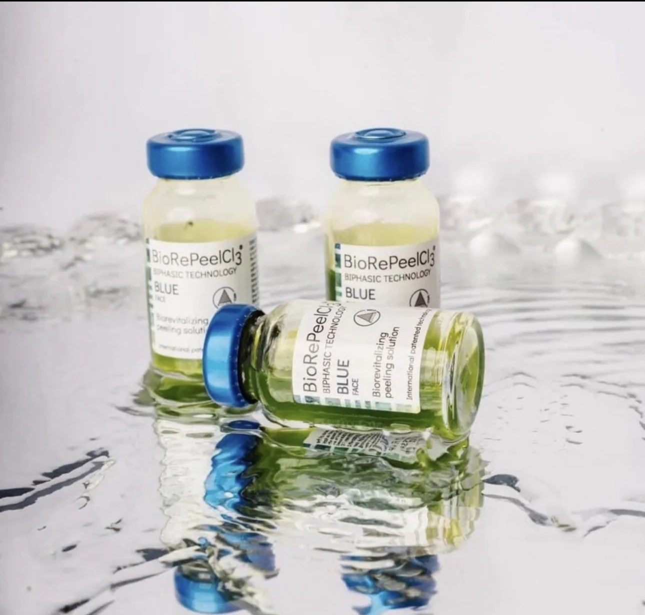 Three vials of BioRePeelCl3 with blue caps on a reflective wet surface, two upright and one lying down, containing a yellow-green solution.