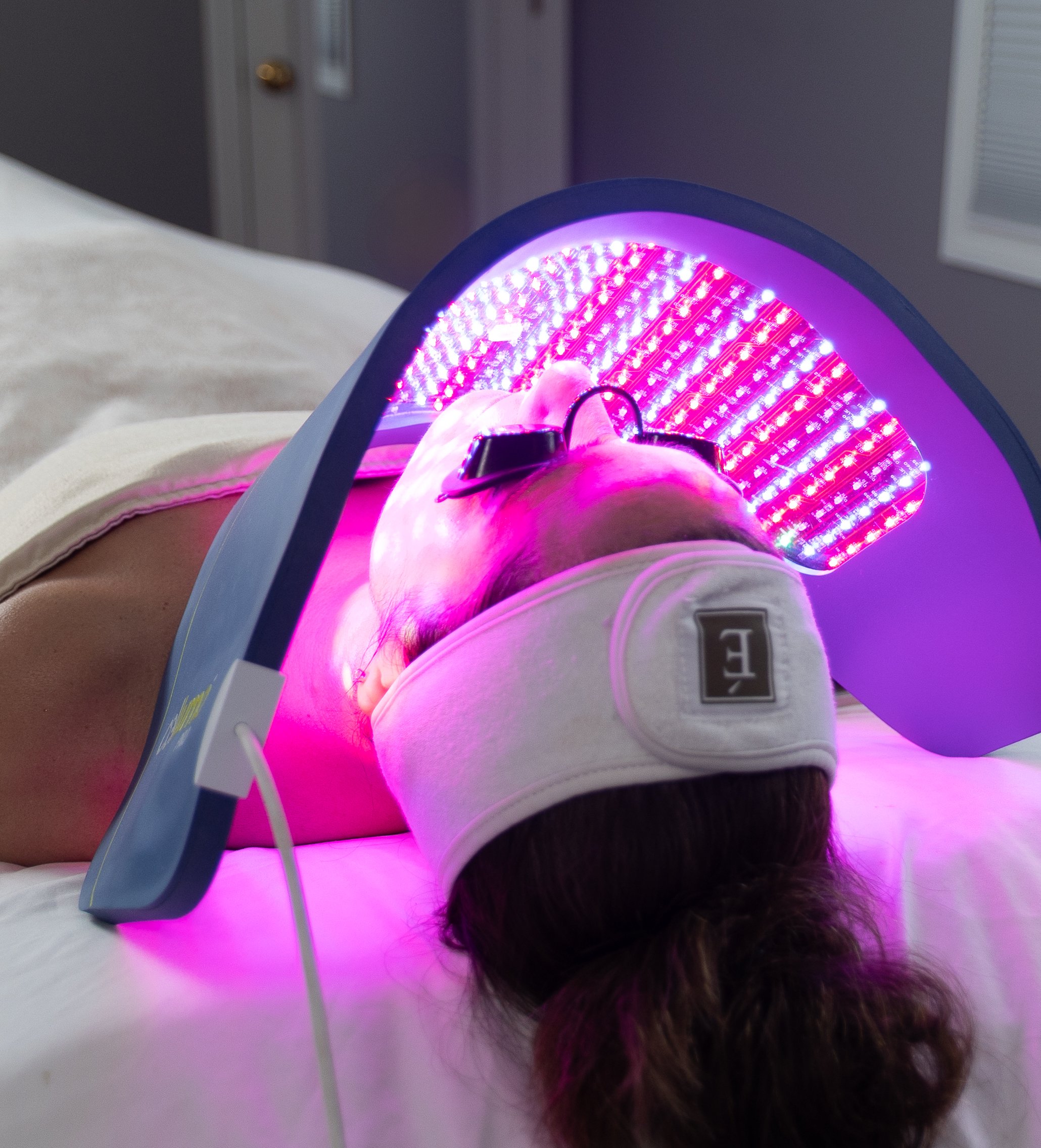 A person lying on a bed undergoing a facial treatment with LED light therapy, wearing protective glasses and a white headband, with purple and pink LED lights illuminating their face.