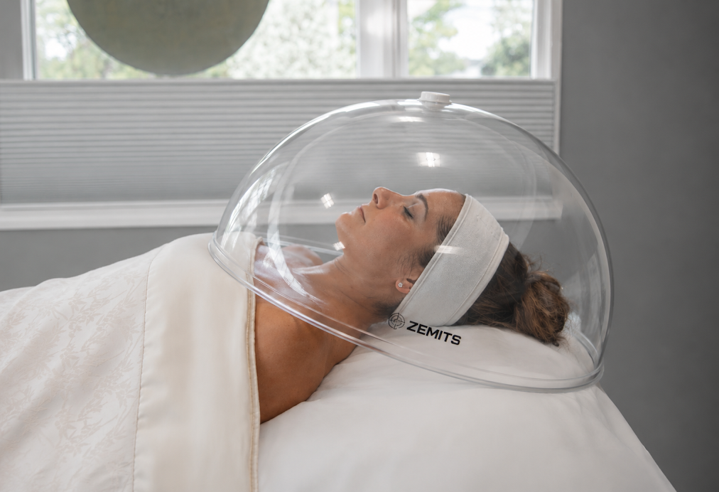 A woman lying on a medical bed with her head inside a clear protective helmet.