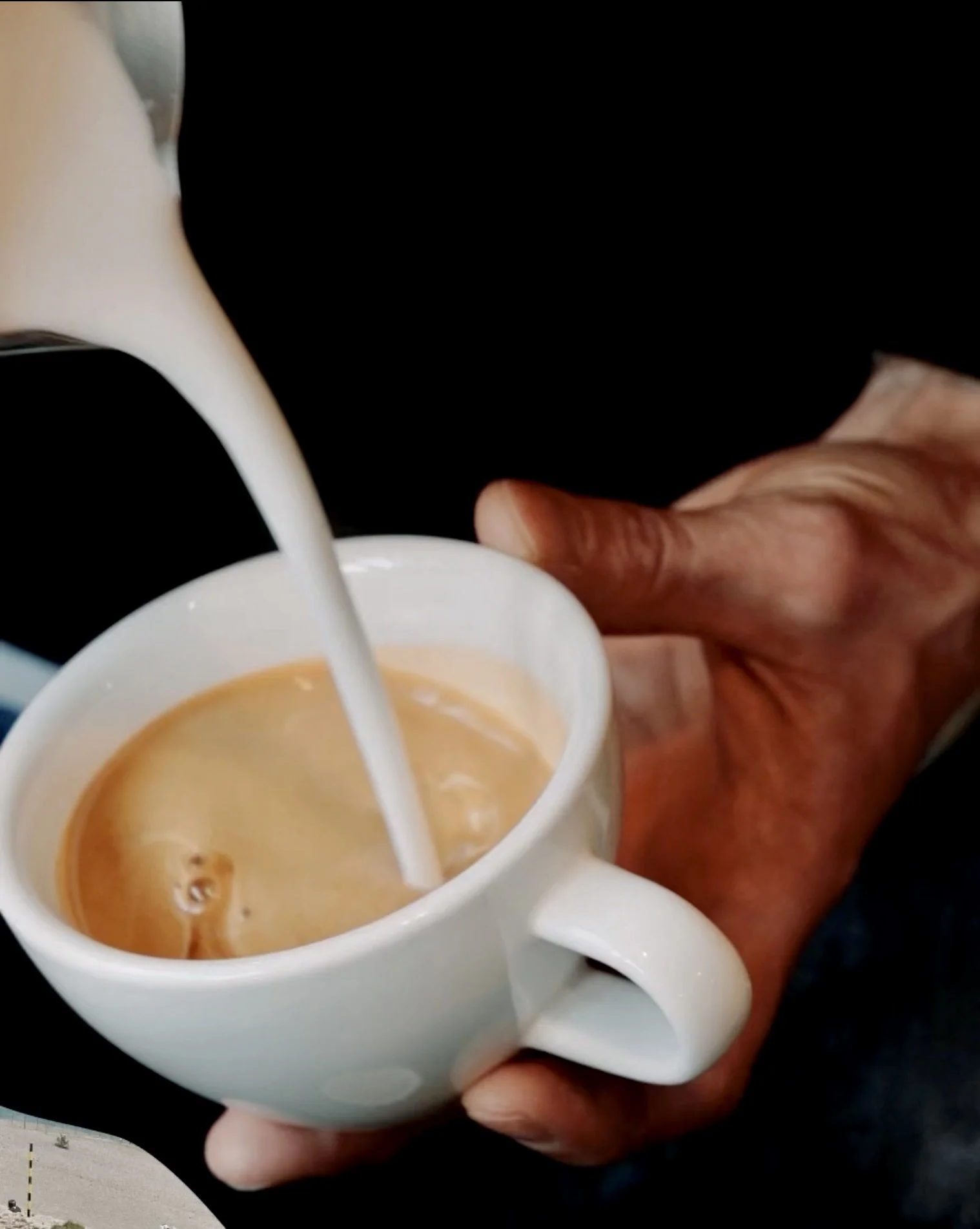 Une main tient une tasse blanche dans laquelle on verse du lait à café ou un café au lait, créant une infusion de couleurs marron clair.