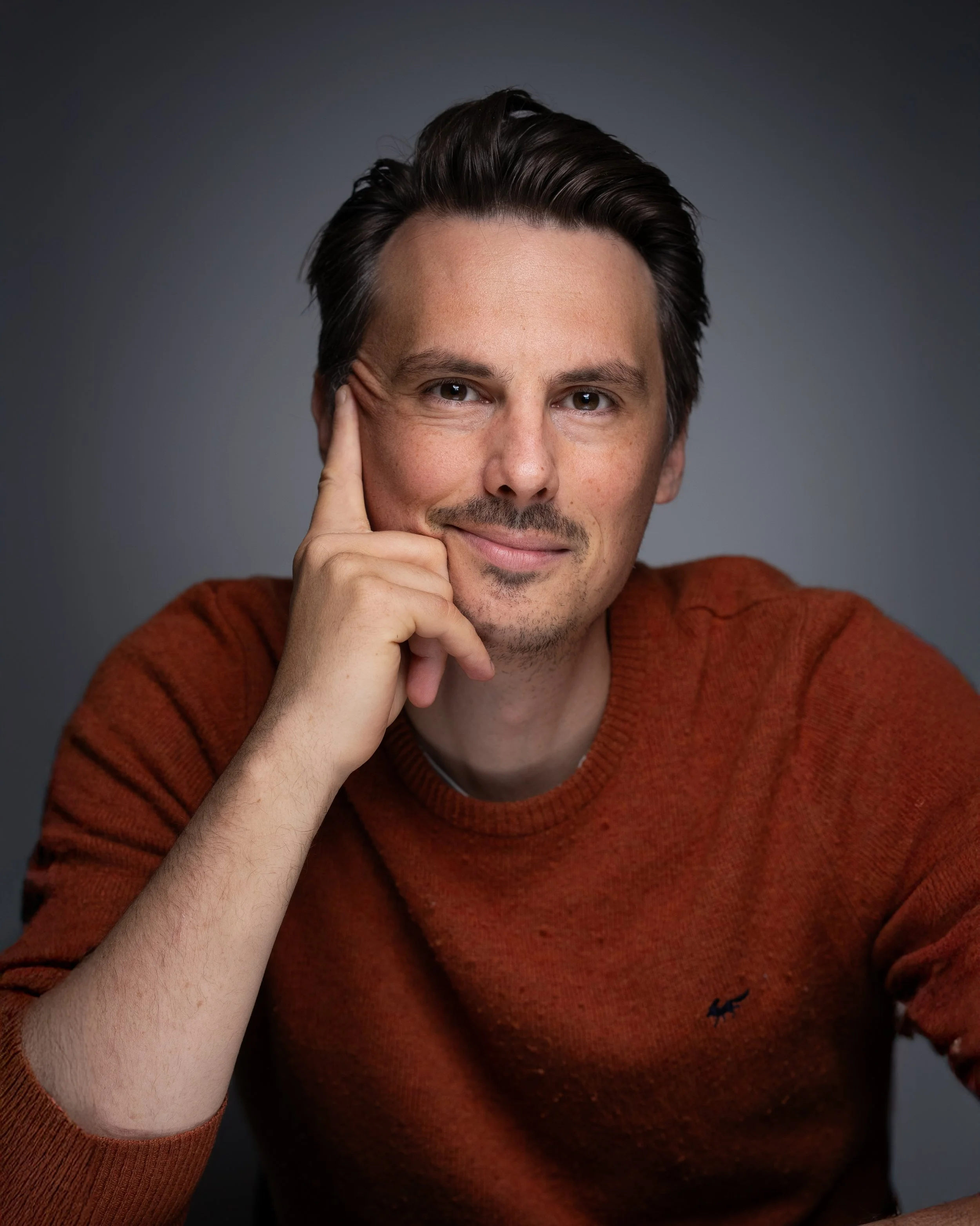 Portrait of a man with dark hair and a slight smile, wearing an orange sweater, resting his head on his hand against a dark gray background.