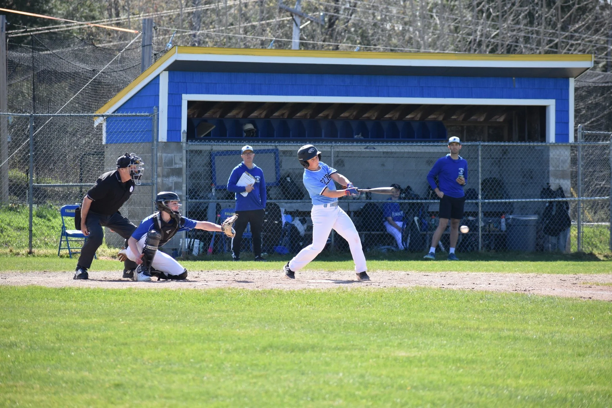 A youth baseball player swings at a pitch during a game, with the catcher and umpire crouched behind him, and coaches and teammates watching from the dugout.