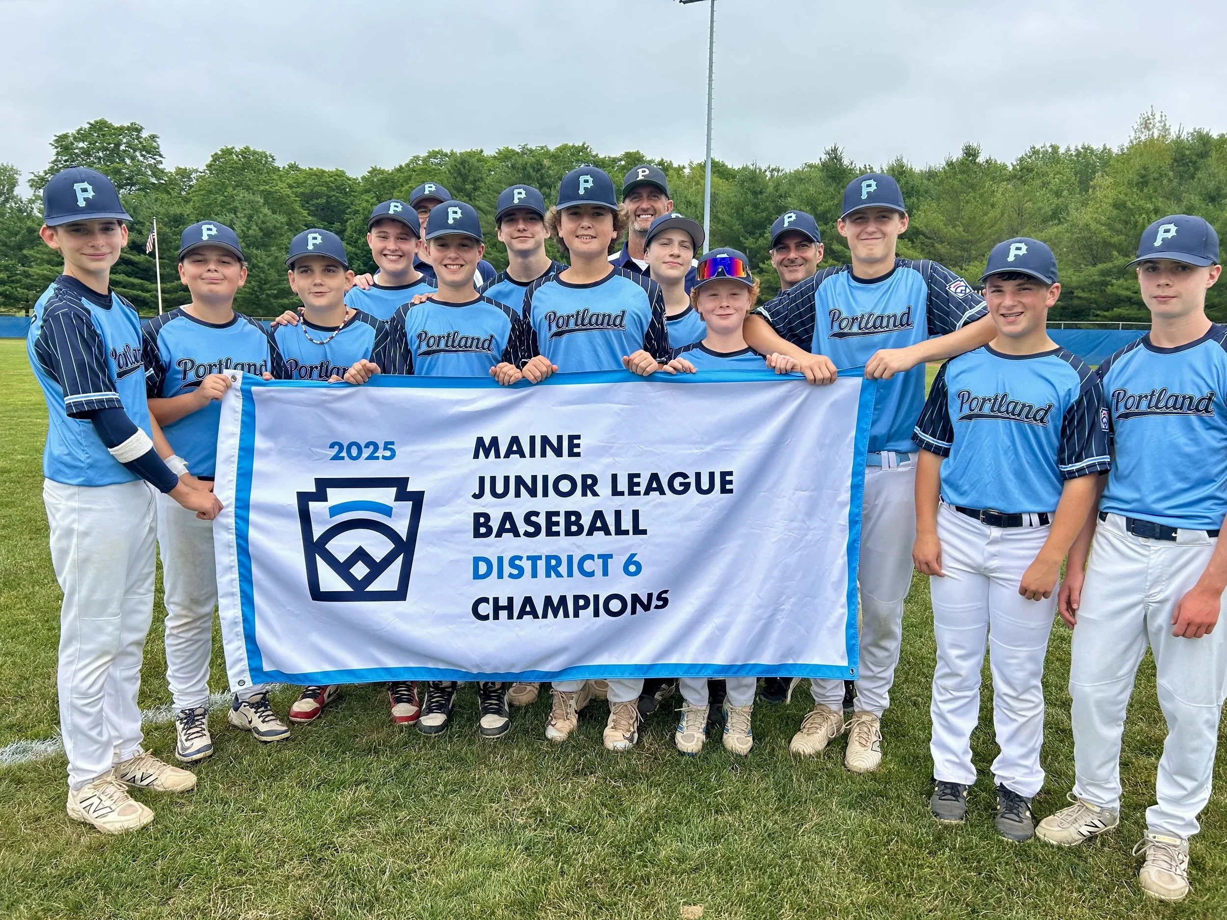 Youth baseball team posing on a field holding a banner that reads 2025 Maine Junior League Baseball District 6 Champions, wearing blue and white uniforms with caps, some team members have pink shoes, and a few are wearing sunglasses, with trees and a cloudy sky in the background.
