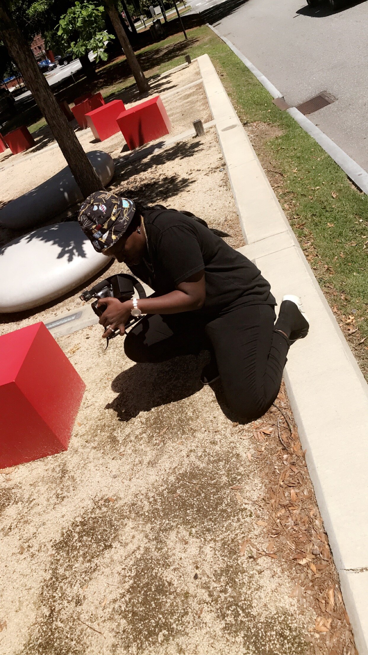 A woman kneeling on the ground in a park, holding a camera and looking down at the ground. She is wearing a black outfit, a patterned bucket hat, and sunglasses. The park has trees, red benches, large black spherical objects, and a sandy area with a 
