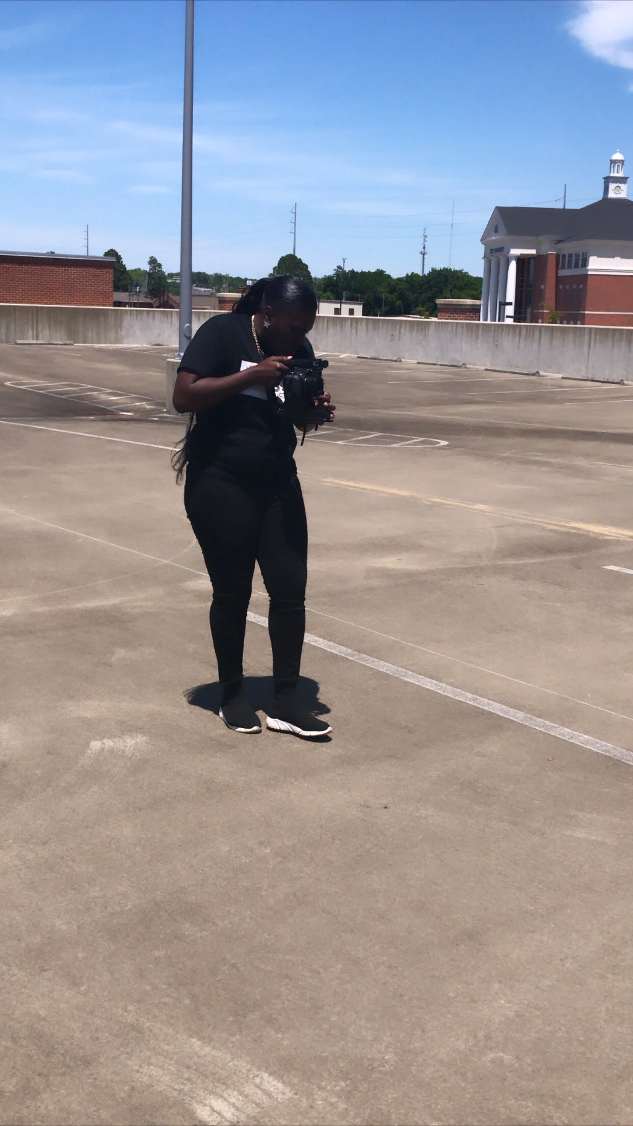 A woman in black clothing holding a camera in an empty parking garage under a clear blue sky.