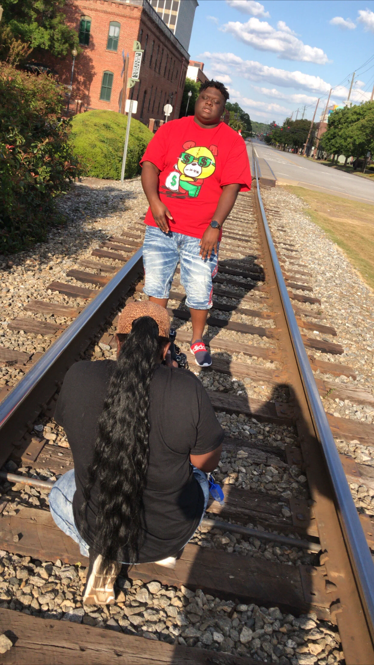 A man standing on railroad tracks with a photographer crouched down taking pictures of him. The man is wearing a red graphic t-shirt, denim shorts, and sneakers. The background shows a street with shops, trees, power lines, and a partly cloudy sky.