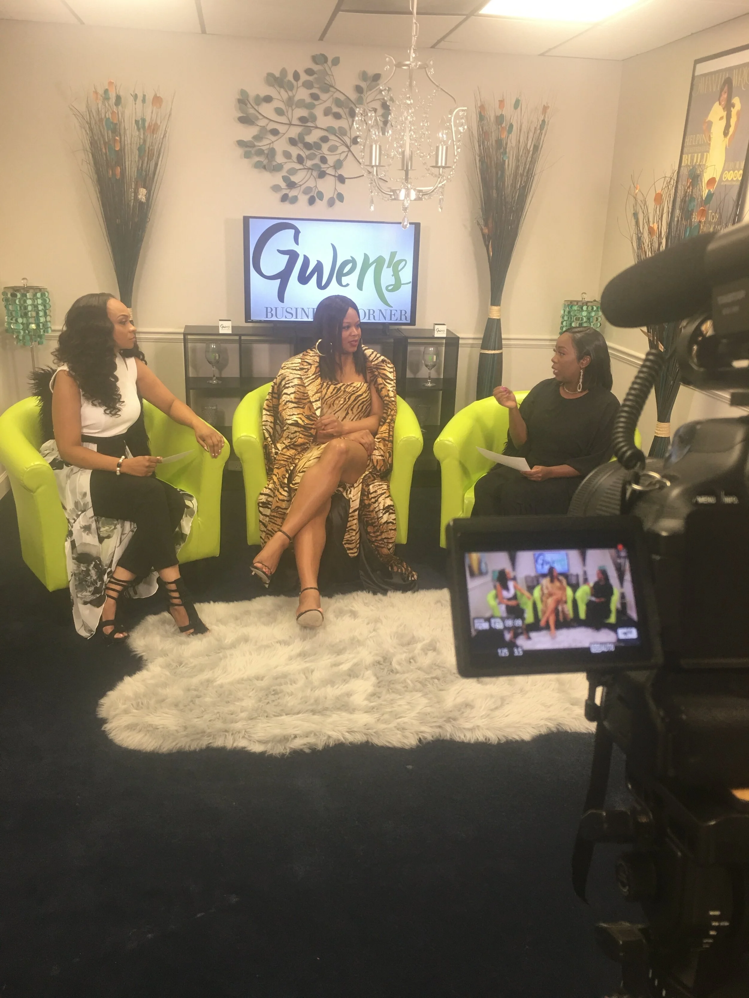 Three women are seated on bright green chairs in a TV studio set, engaged in conversation. Behind them is a sign reading 'Gwen's Business Blender' on a television screen. The set is decorated with tall vases of sticks and flowers, a chandelier, and a