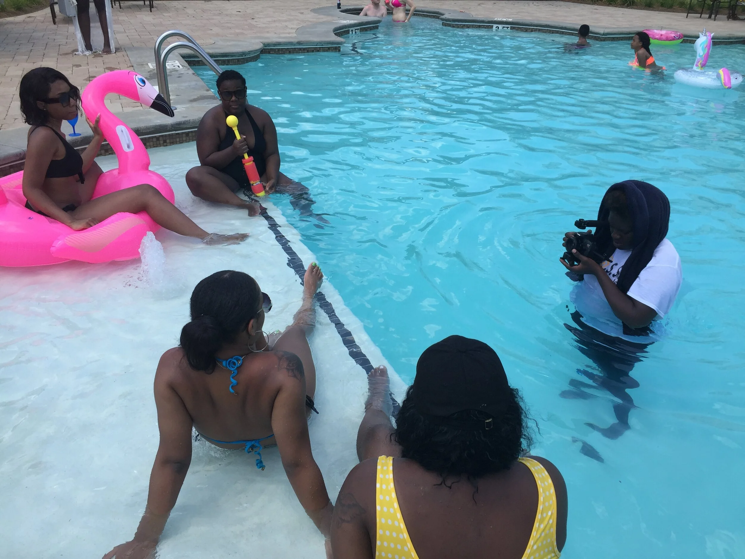 Group of people sitting and standing near a swimming pool, some in the water, one person holding a camera, with inflatable pool floats, including a pink flamingo float, in the scene.