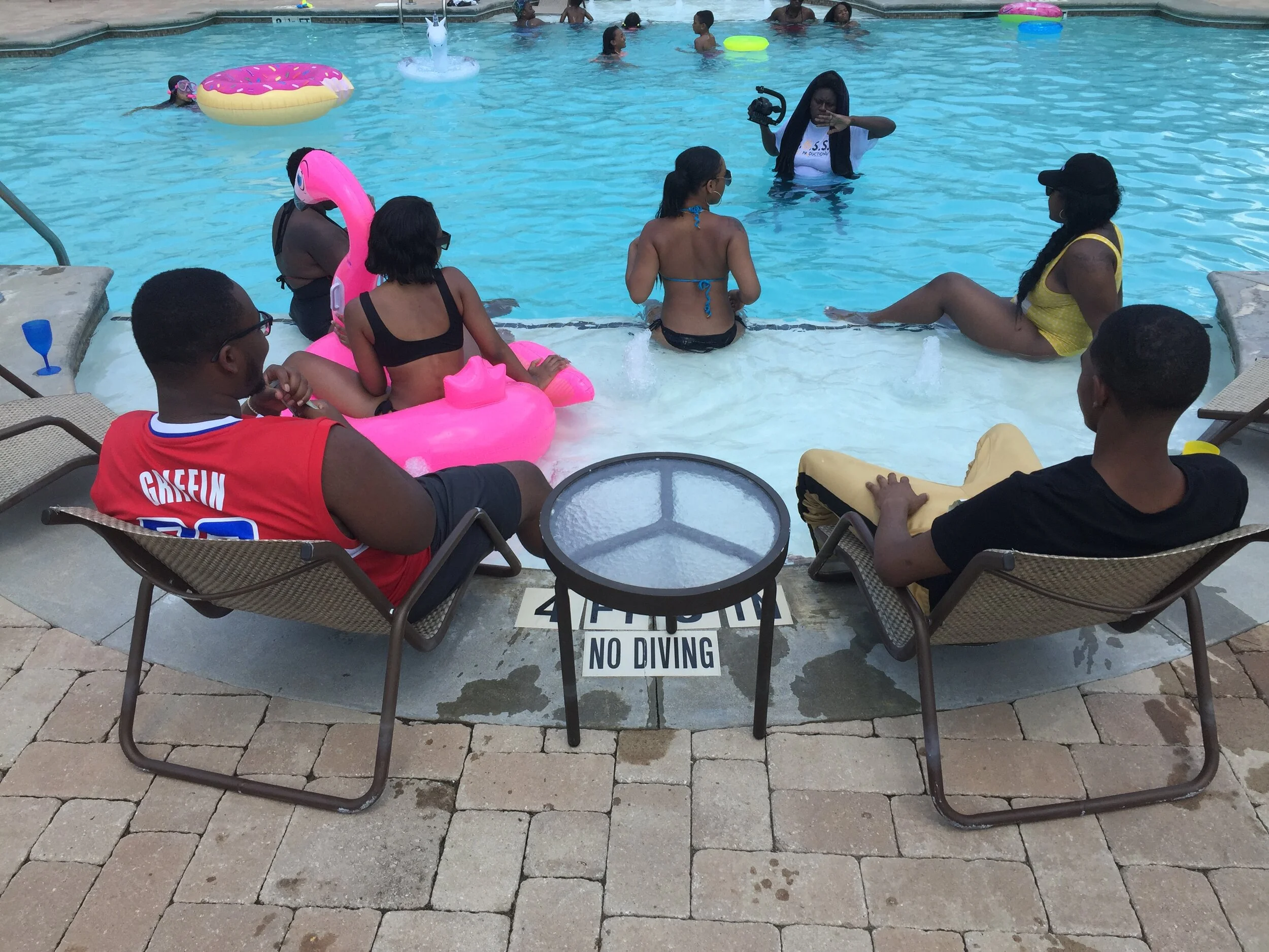 People relaxing by a swimming pool, some sitting on lounge chairs and others in the pool, with flotation devices and a woman filming with a camera.