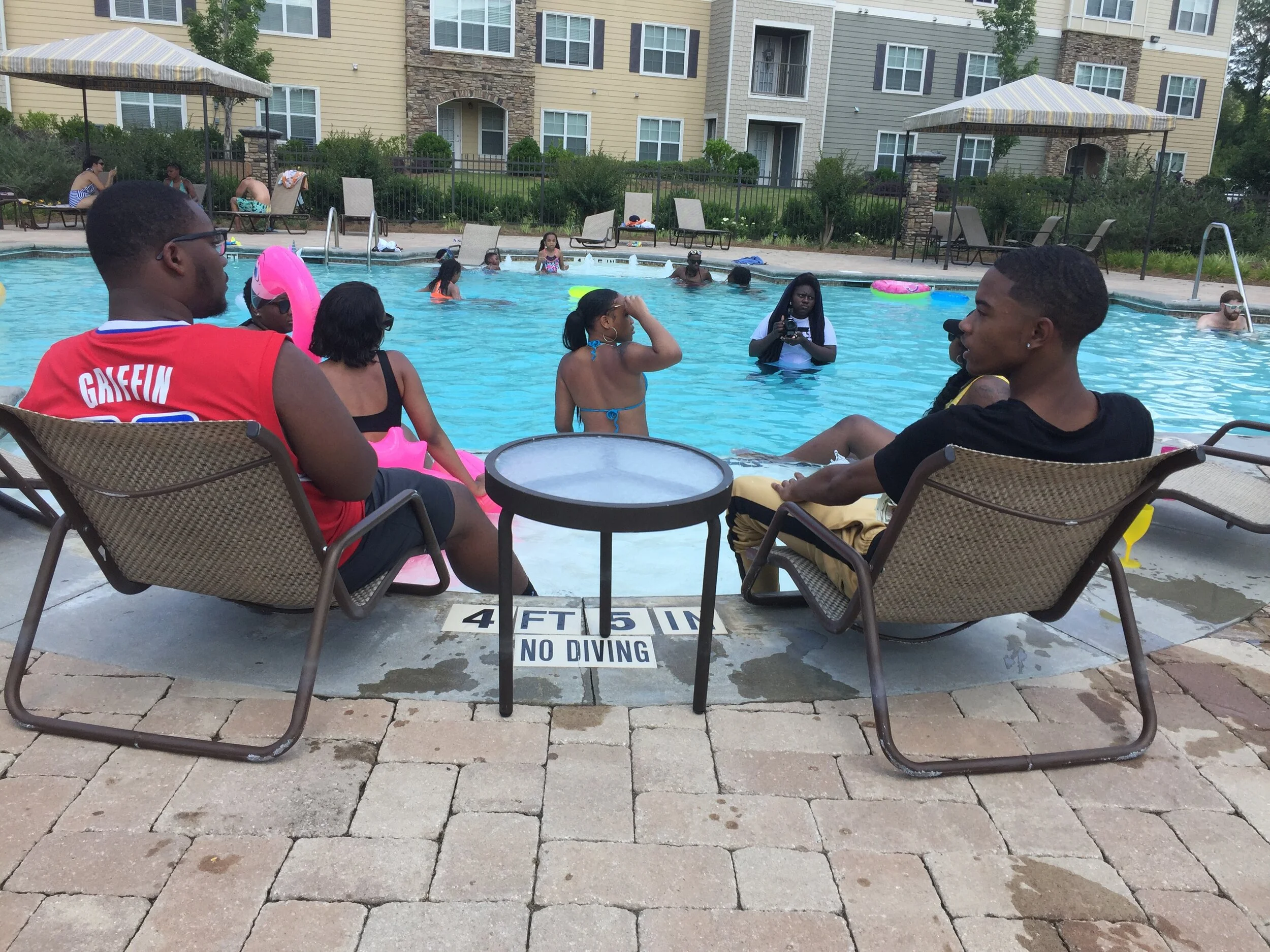 People relaxing by a swimming pool, some sitting on lounge chairs and others swimming or playing in the water. There are buildings and umbrellas in the background.