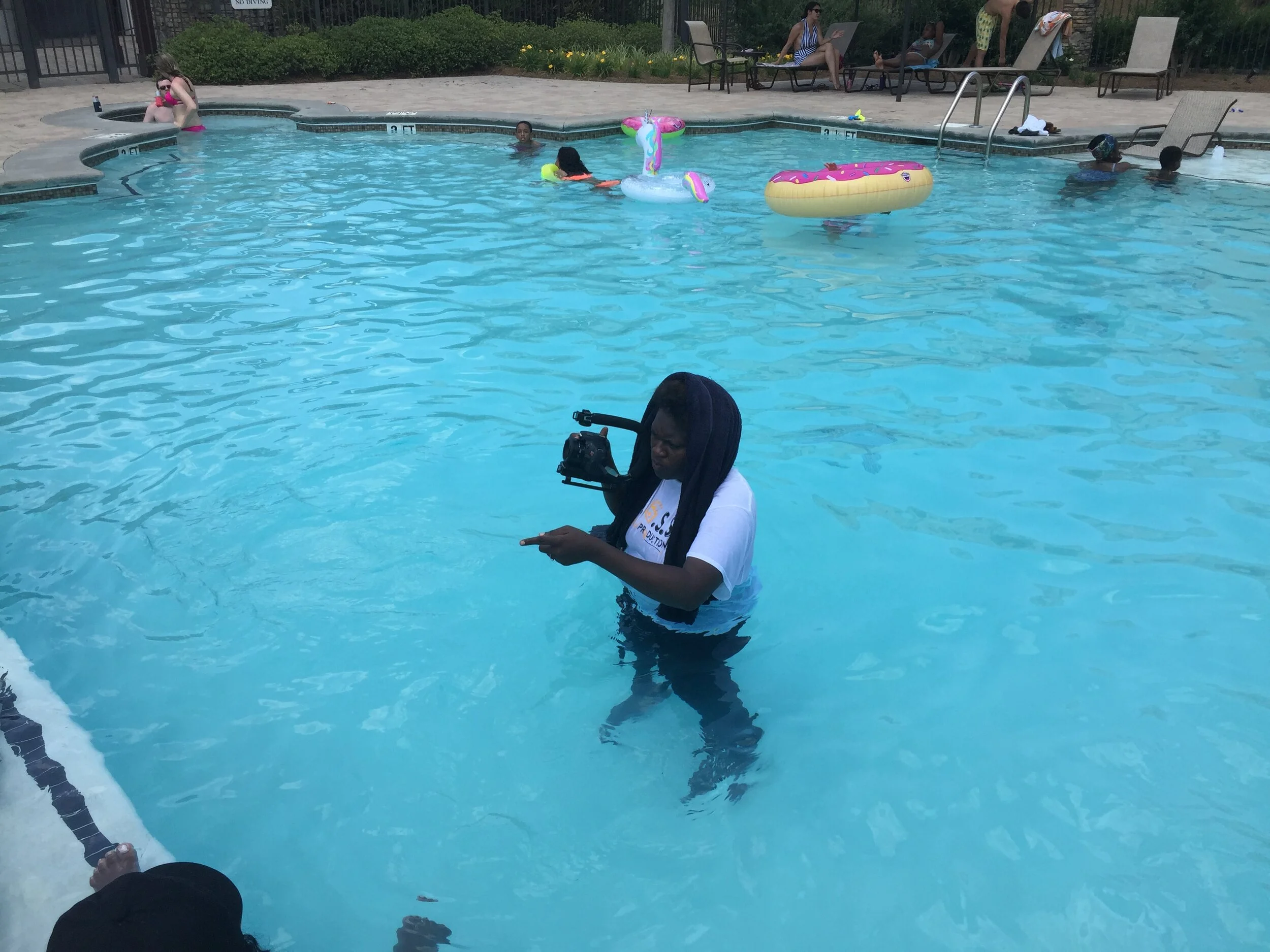 Person standing in a swimming pool with underwater camera mounted on shoulder, while other people swim and relax around the pool.