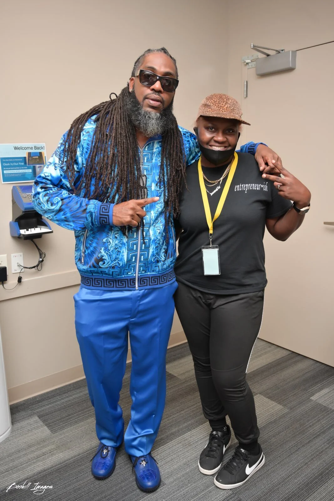 Two people posing together indoors, one man wearing blue patterned jacket and pants, sunglasses, and the other woman in black T-shirt with 'entrepreneur' written on it, black pants, sneakers, and wearing a hat. They are smiling and making hand gestur