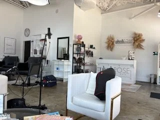 Inside a salon or studio with a white reception desk, a white armchair with a black cloth, and various equipment including a camera on a tripod, mirrors, and shelves with products.