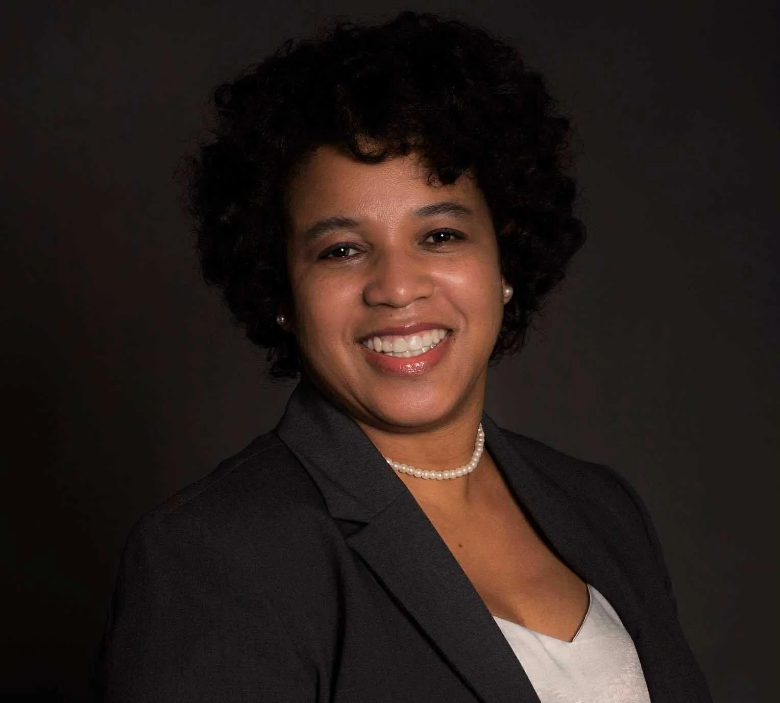 Portrait of a woman with curly black hair, smiling, wearing a dark blazer, pearl necklace, and earrings, against a black background.
