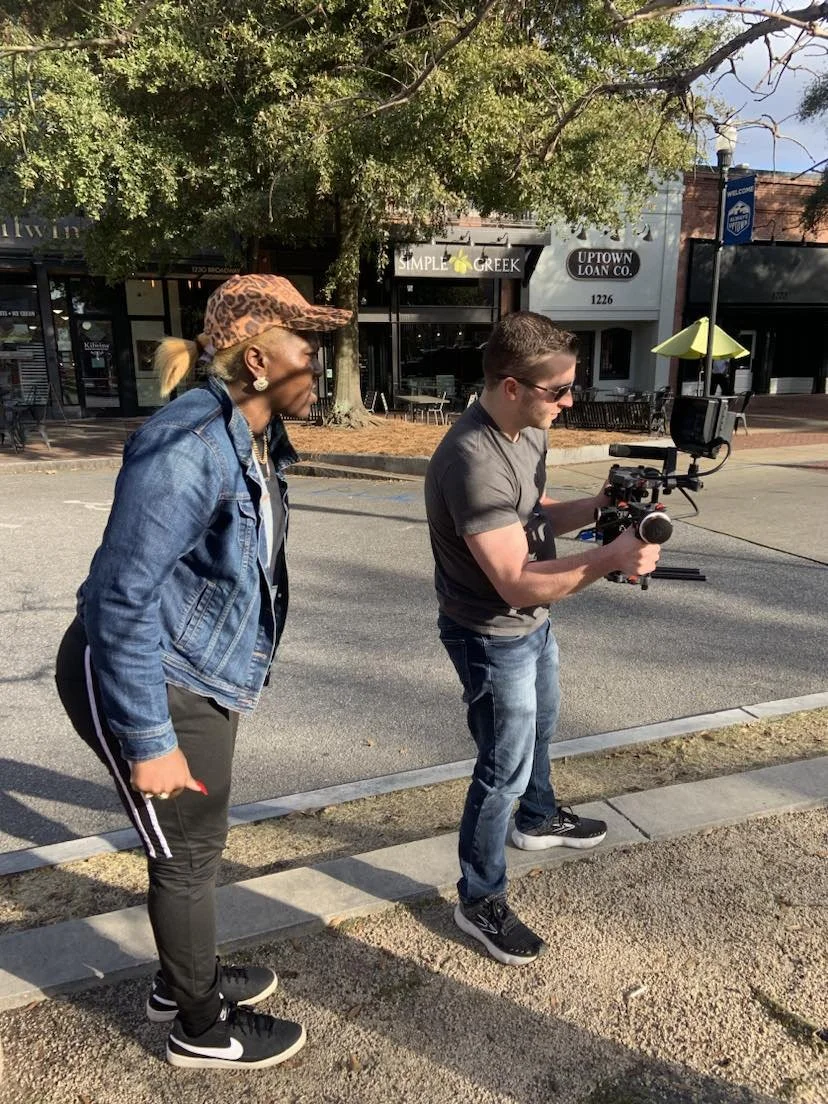 A woman and a man are standing on a sidewalk with the woman watching as the man operates a camera stabilizer. The woman is wearing a leopard print cap, denim jacket, dark pants with white stripes, and Nike sneakers. The man is wearing sunglasses, a g