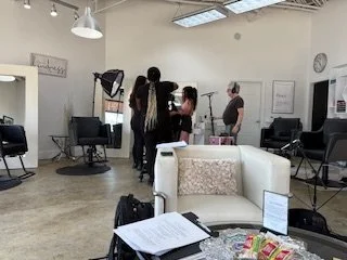 A group of people gathered in a hair salon or studio with styling chairs, mirrors, and lighting equipment.
