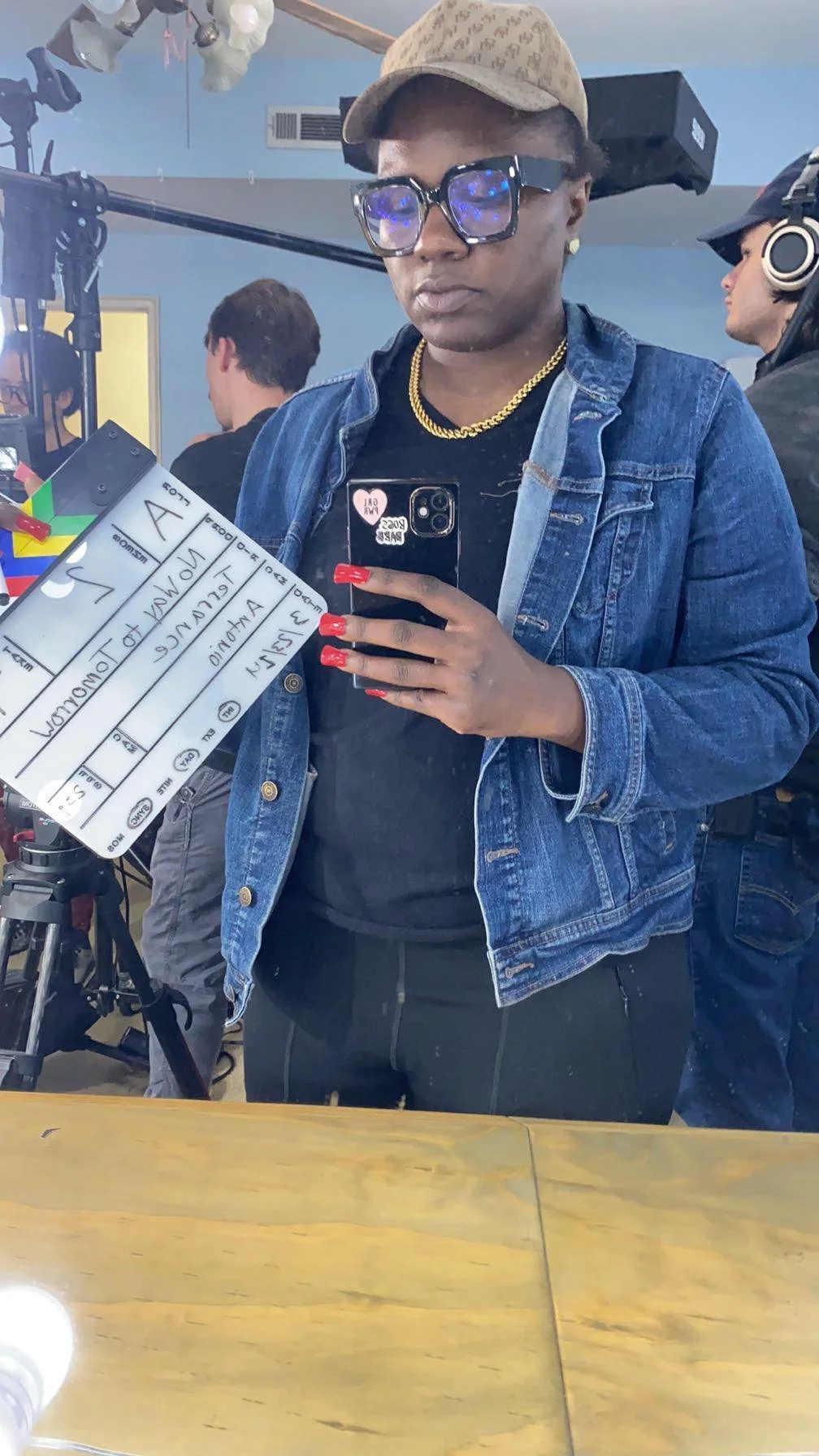 A woman in a denim jacket taking a selfie with her phone, standing in front of a mirror. She is wearing glasses, a beige cap, gold earrings, a gold necklace, and has red painted nails. The setting appears to be a film or photo shoot, with crew member