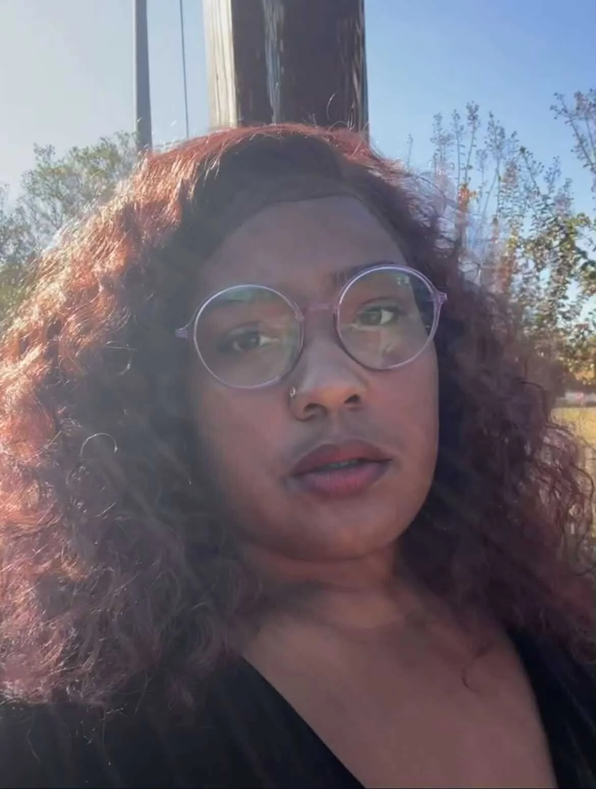 Close-up portrait of a woman with curly hair, wearing round glasses, outdoors on a sunny day with trees and a building in the background.