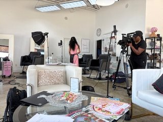 A behind-the-scenes filming or photo shoot setup in a room with a woman, camera operator, and various equipment, including lighting, chairs, and a table with magazines.
