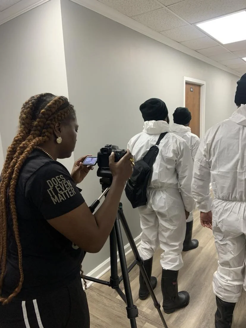 A woman with long braids and a black shirt with the text 'Does It Even Matter?' is filming or taking pictures of people in white protective suits walking in a hallway.