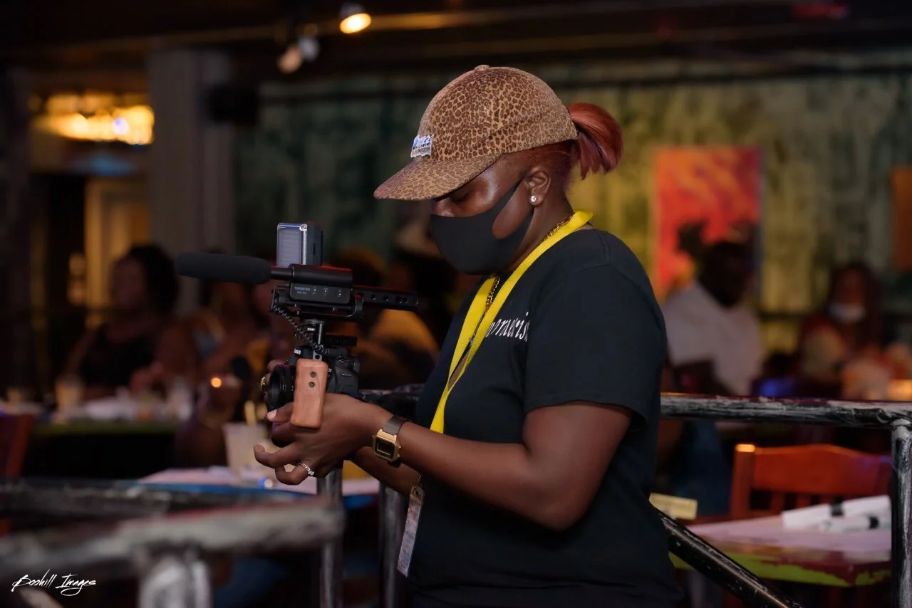A woman wearing a leopard print cap, black face mask, and black shirt operating a camera on a stabilizer inside a dimly lit restaurant or café. She is focused on adjusting the camera, with several people seated in the background.