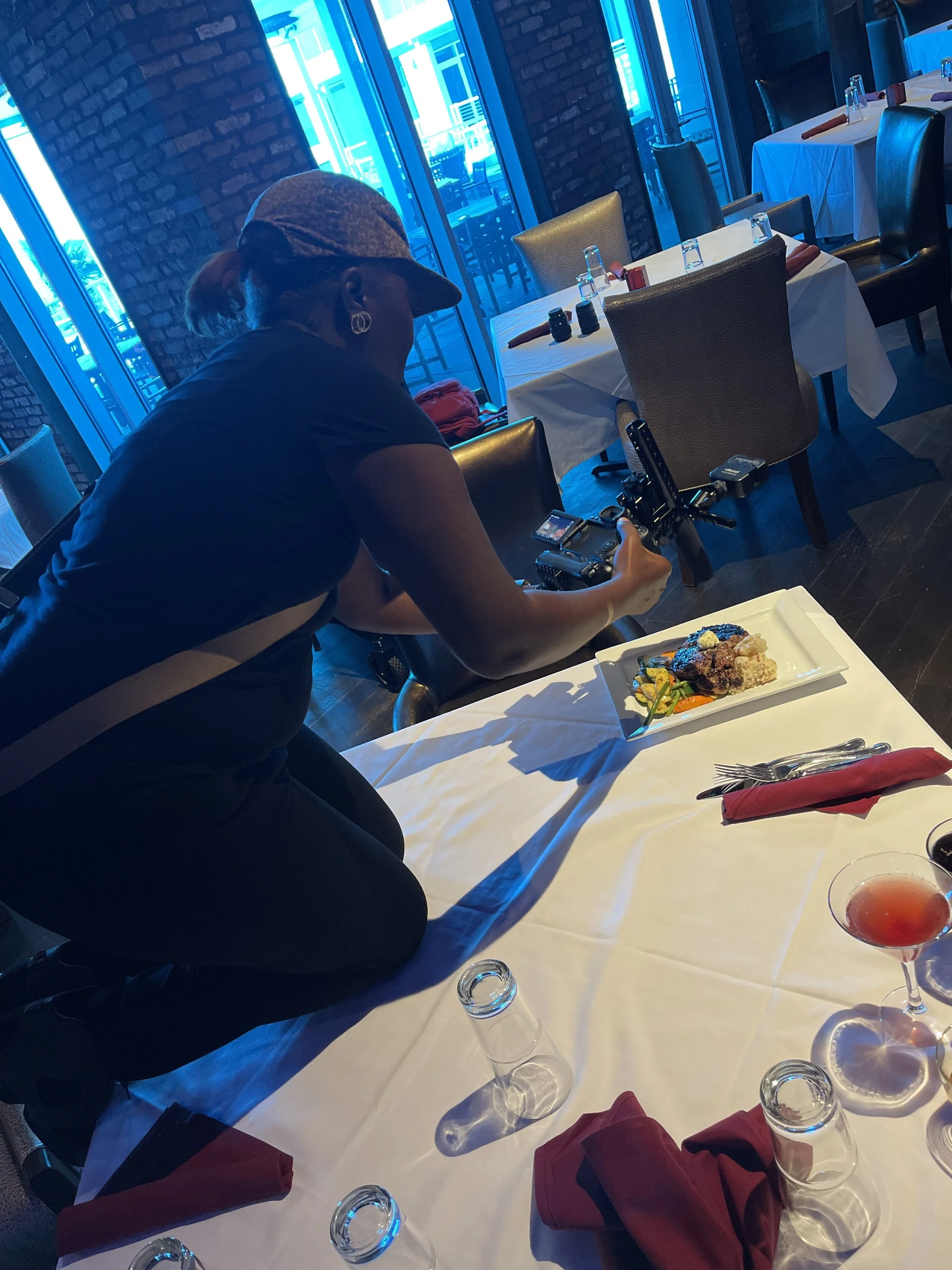 Woman in black playing camera at an elegant dining table with white tablecloth, wine glasses, and red napkins, in a restaurant with large windows and brick walls.