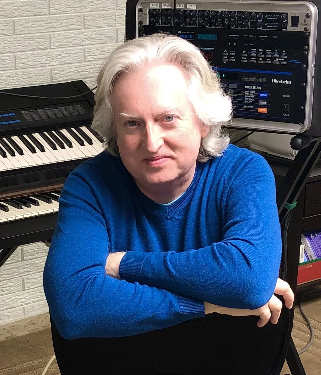 A man with shoulder-length gray hair, wearing a blue sweater, sitting in front of electronic music equipment including a keyboard and a synthesizer.