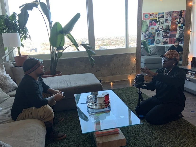 Two men are in a living room with large windows, one sitting on a sofa with a gaming controller, and the other kneeling with a camera, engaging in an interview. The room has houseplants, a collage of colorful artwork on the wall, and string lights.