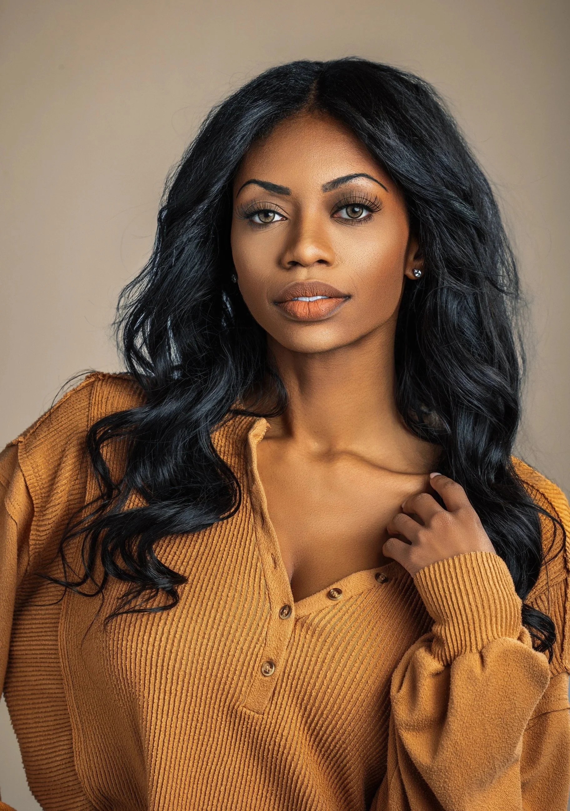 A woman with long wavy black hair and makeup, wearing a brown button-up sweater, looking at the camera.