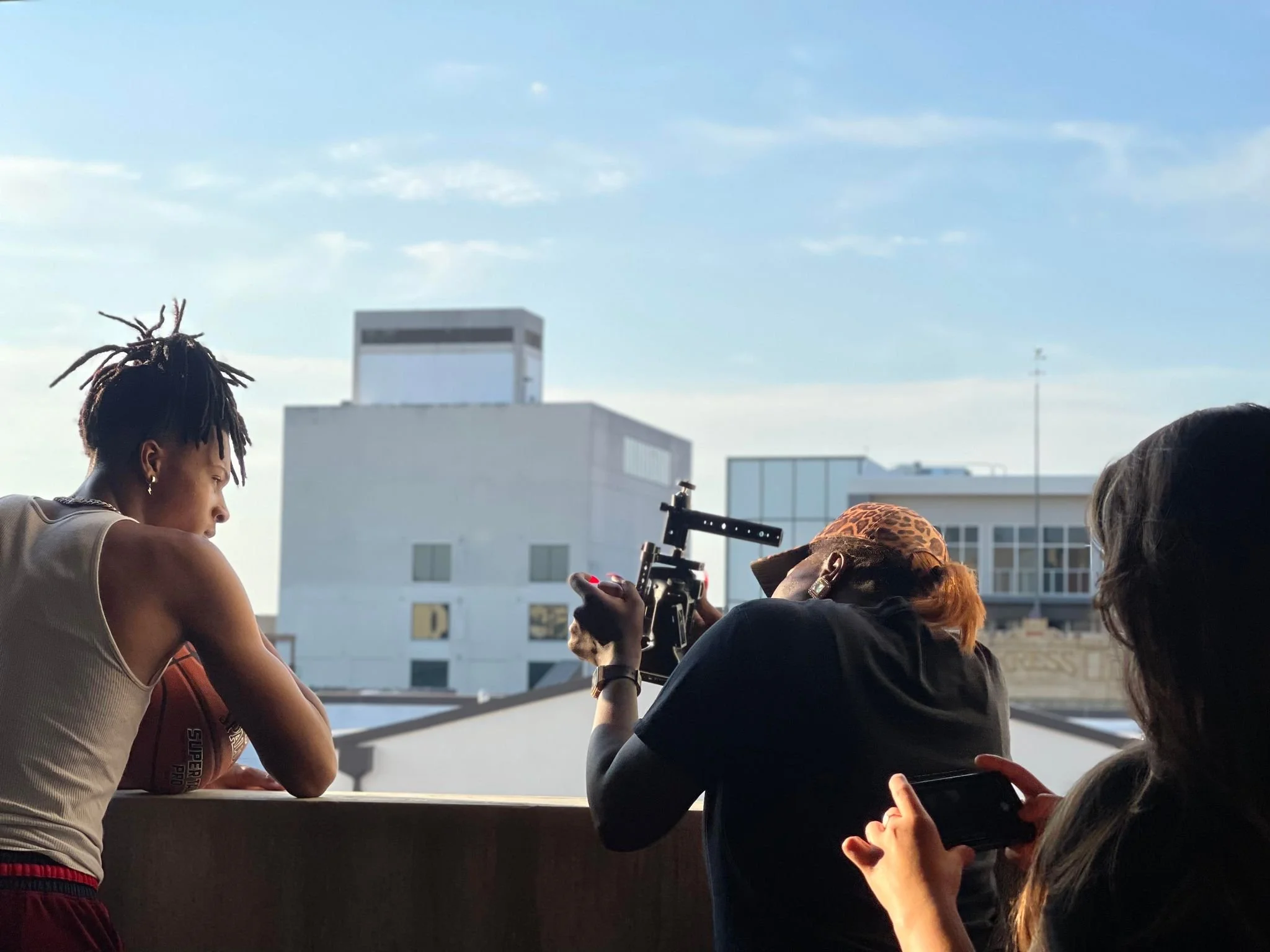 Three women on a rooftop, one in a tank top holding a basketball, another with red hair and an orange headband aiming a film camera, and the third woman with long dark hair taking a photo with a smartphone, with city buildings in the background.