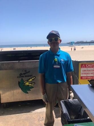 Un hombre sonríe frente a una parrilla de asado de maíz en una playa.