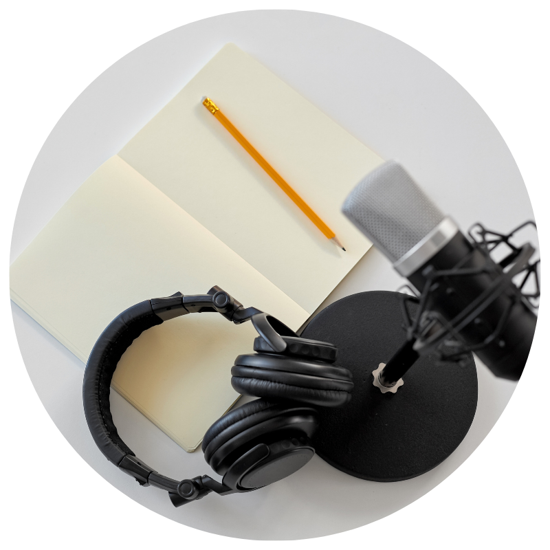 A microphone with a pop filter, a pair of black headphones, an open notebook, and an orange pencil on a white surface.