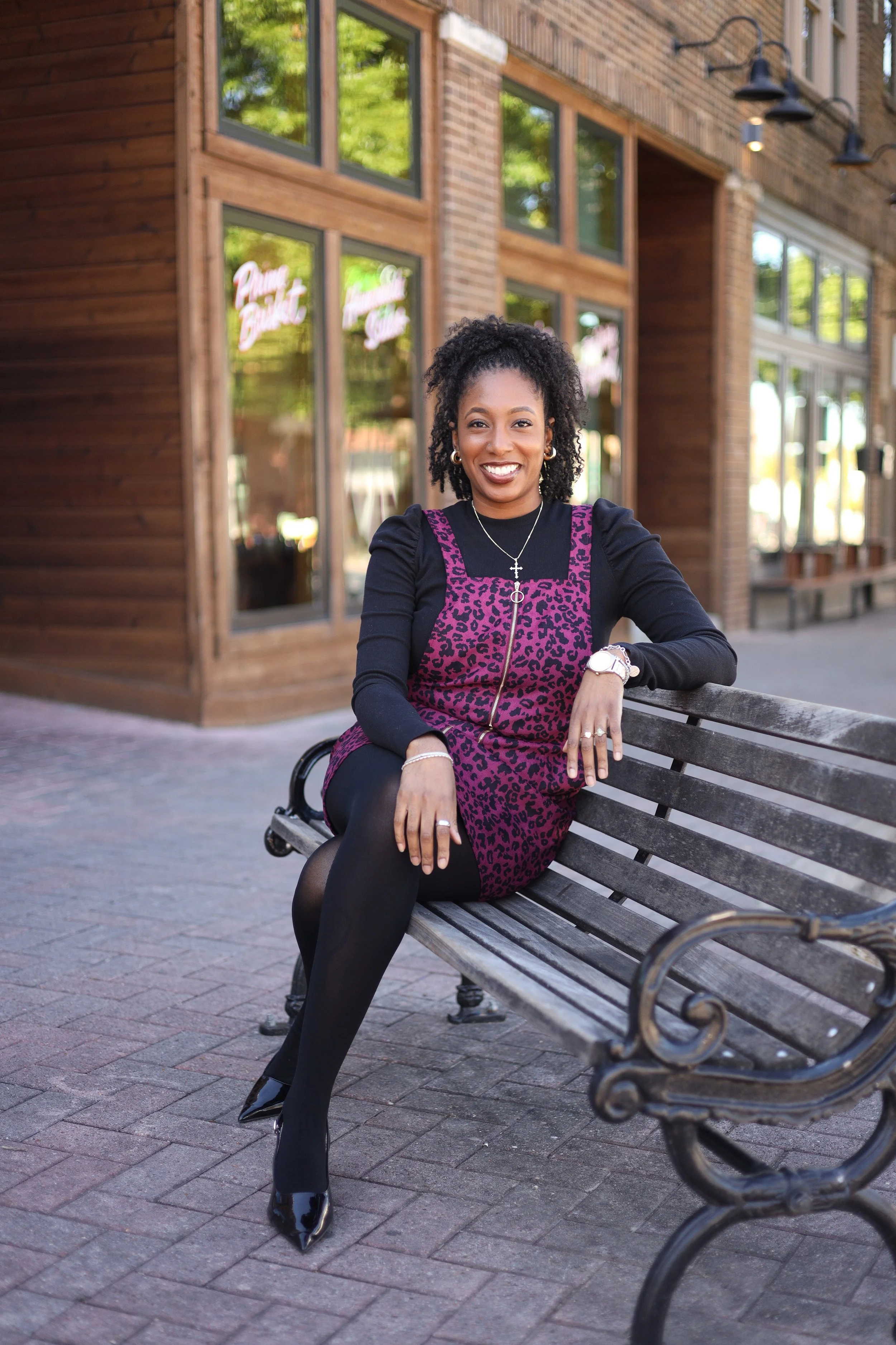 A woman with curly hair, wearing a black long-sleeve shirt and leopard print purple dress, is sitting on a park bench outside a brick building with large windows. She is smiling, showing her teeth, and wearing a necklace, watch, rings, and black tights with high heels.