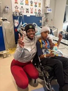 A young girl in a hospital room, smiling and making a peace sign, sitting next to a child with medical equipment, both appearing positive.