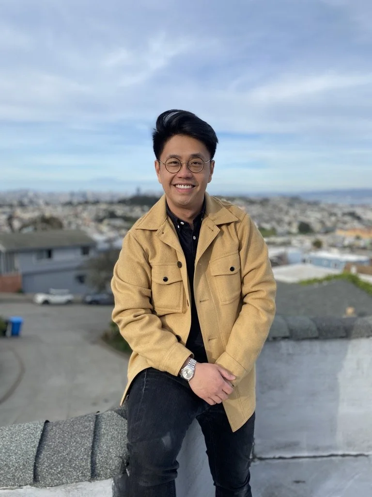 Young man with black hair, glasses, wearing a tan jacket and black shirt, smiling while leaning on a rooftop ledge, with a cityscape and blue sky in the background.