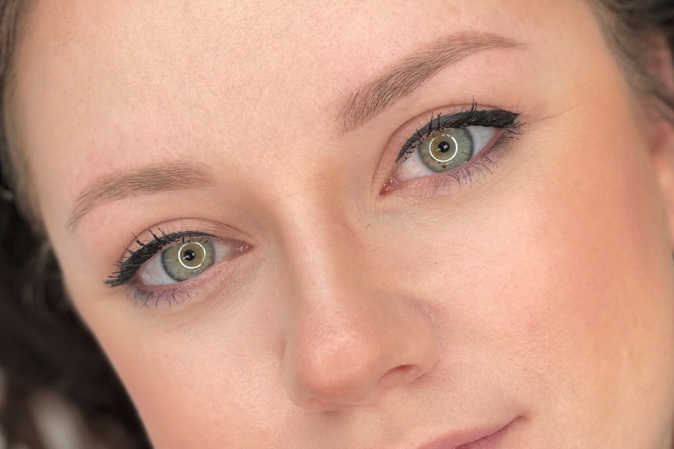Close-up of a woman's face with green eyes, wearing black eyeliner, showing smooth skin and well-groomed eyebrows.