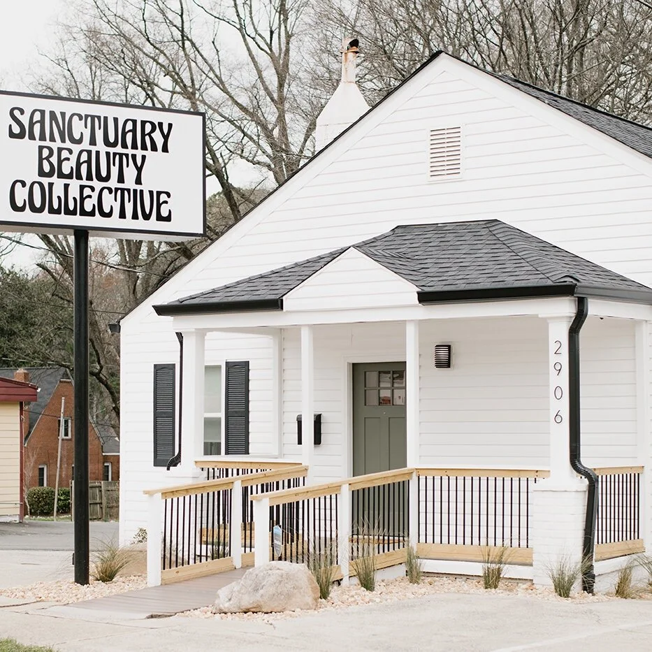 A white building with a sign that reads 'Sanctuary Beauty Collective'. The building has black shutters on the windows, a small front porch with a ramp, and a gray door. There is a rock and some small plants in the front yard.