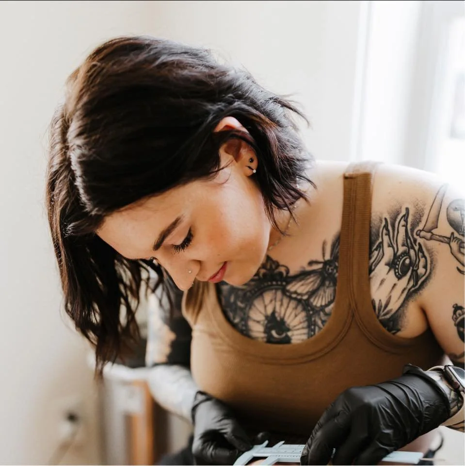 A woman with dark hair, tattoos on her chest and arm, wearing a brown tank top and black gloves, appears to be tattooing or doing some detailed work.