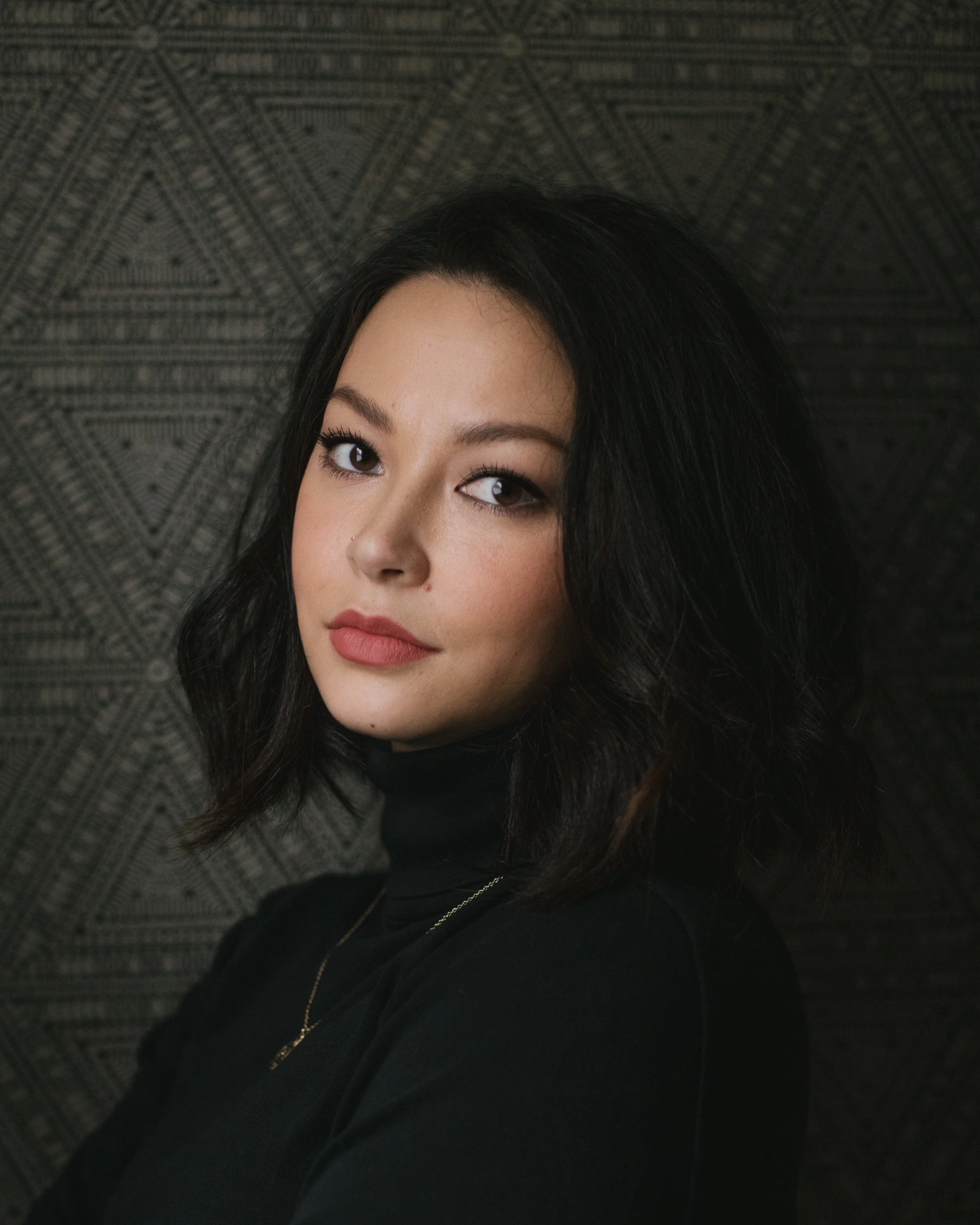 A woman with dark hair, makeup, and a necklace, partially visible against a patterned background.