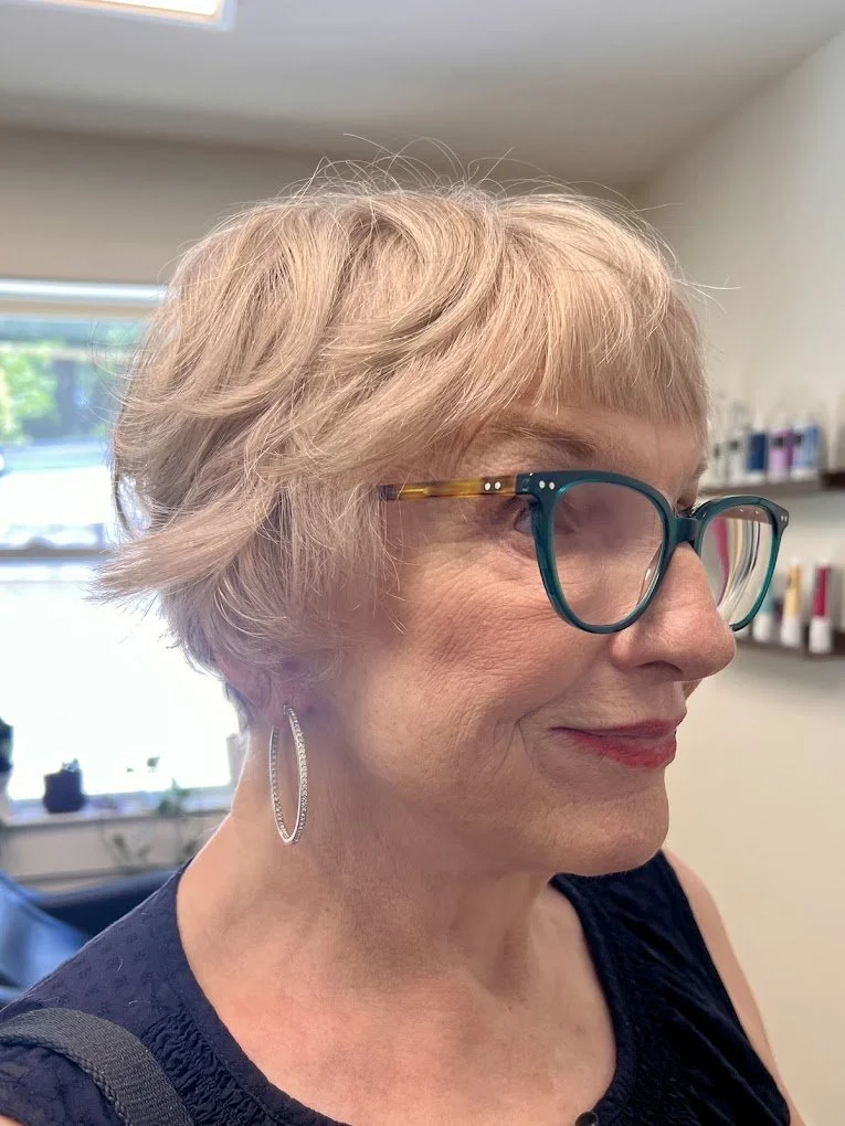A close-up side profile of a smiling older woman with short, wavy blonde hair, wearing teal glasses, large hoop earrings, and a dark blue sleeveless top, indoors with shelves of products in the background.