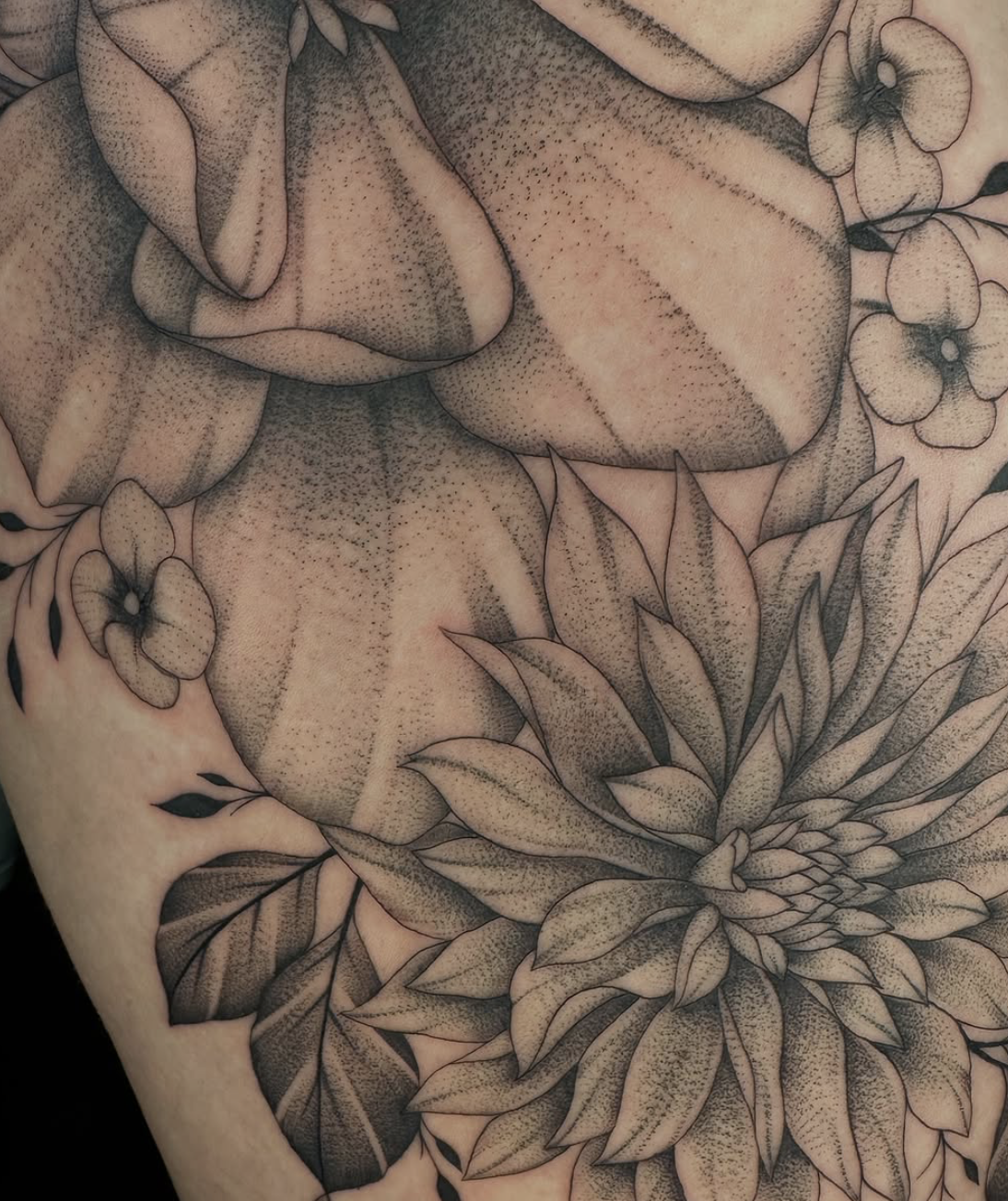 Close-up of a detailed black and gray botanical tattoo featuring a large sunflower, several leaves, and smaller flowers surrounding it.
