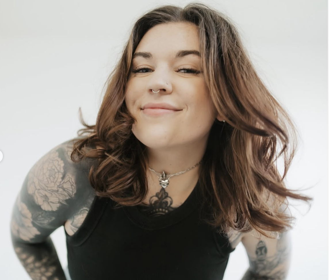 Young woman with wavy brown hair, tattooed arms, and a septum piercing, smiling, wearing a choker necklace and a black sleeveless top, leaning forward.
