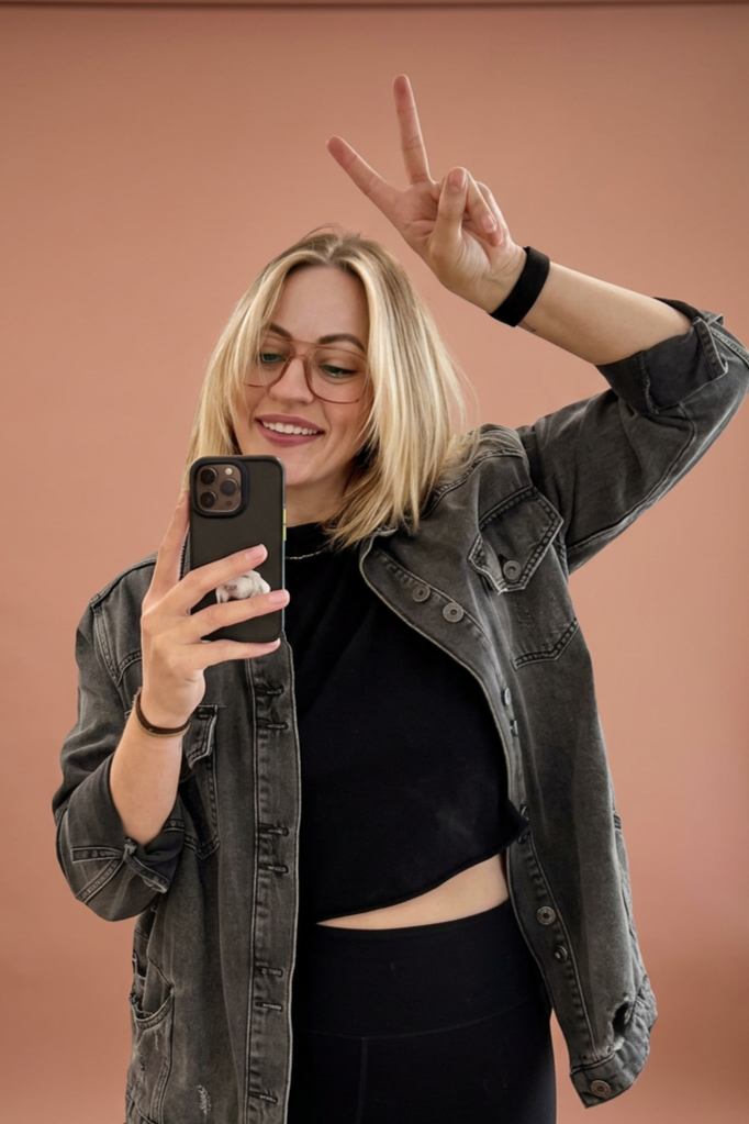 A young woman with blonde hair taking a mirror selfie. She is smiling, making a peace sign with her right hand, wearing glasses, a black t-shirt, black pants, and a gray denim jacket. The background is plain and pink.