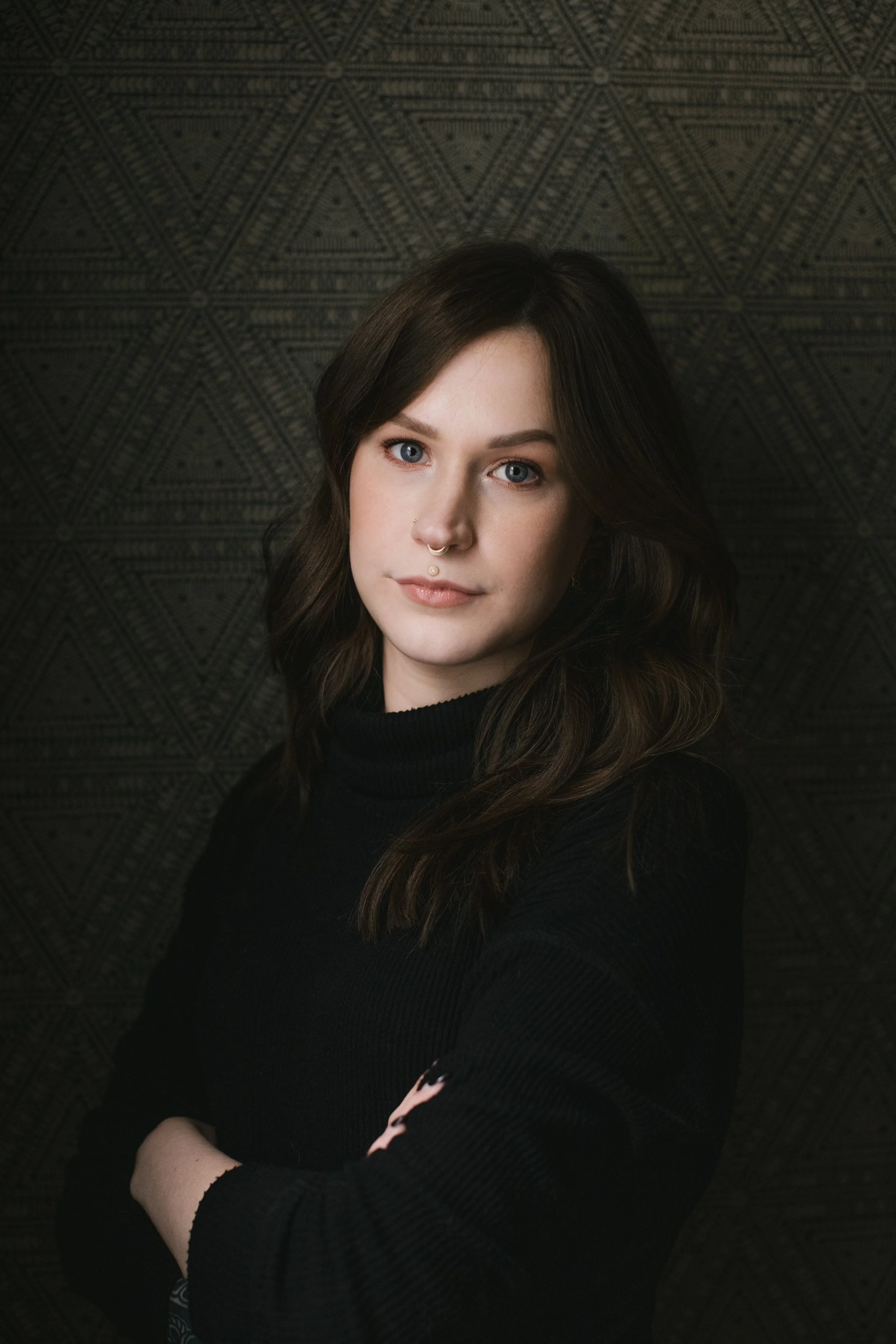 Portrait of a young woman with shoulder-length wavy brown hair, wearing a black turtleneck sweater, standing against a dark geometric patterned background.
