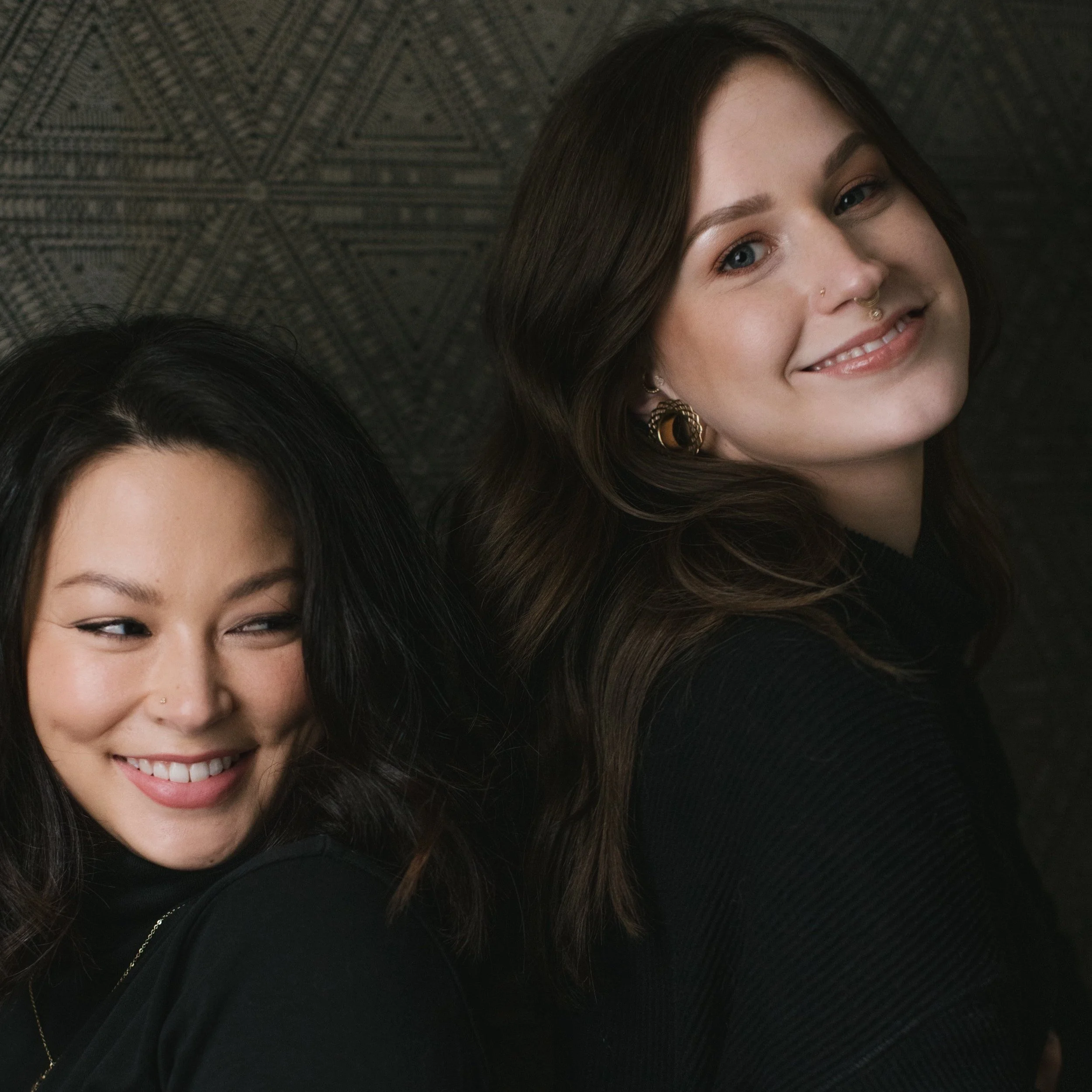Two women smiling, one with dark hair and the other with brown hair, standing close together against a dark patterned background.