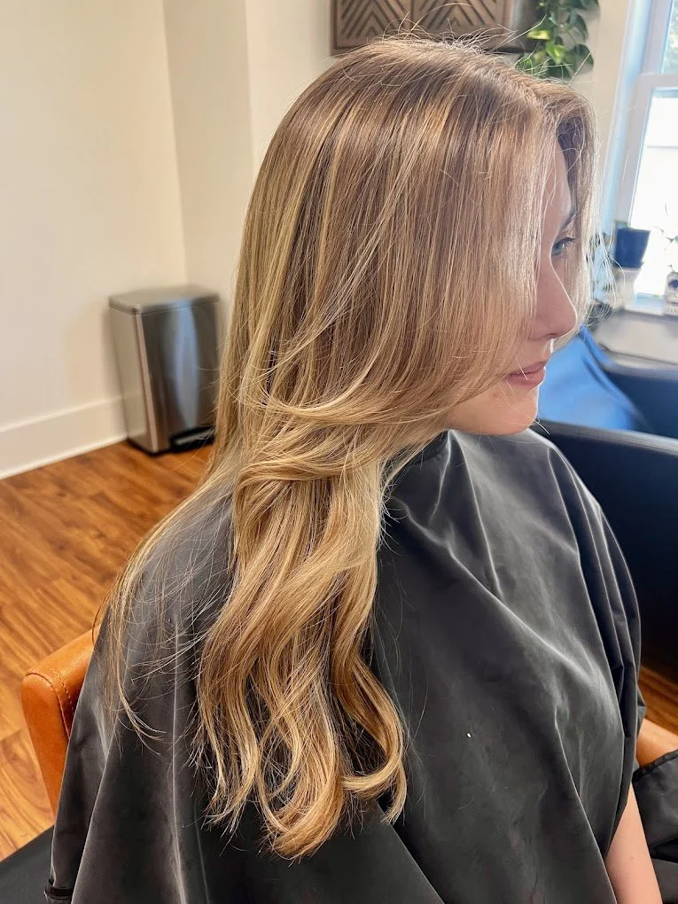 Woman with long, wavy, light brown hair sitting in a salon, wearing a black cape.