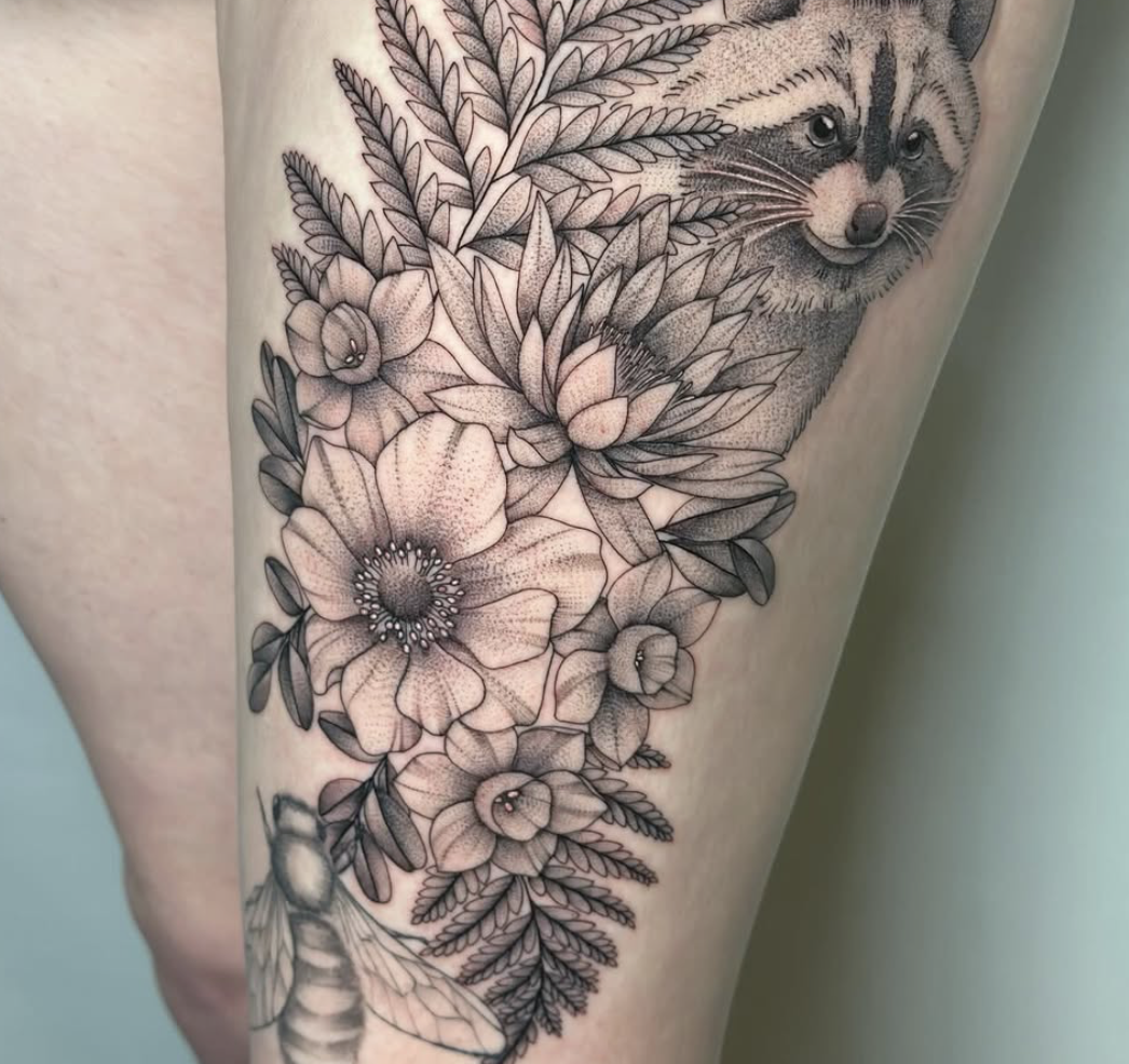 Black and gray floral tattoo with a realistic raccoon and a bee on a person's leg.