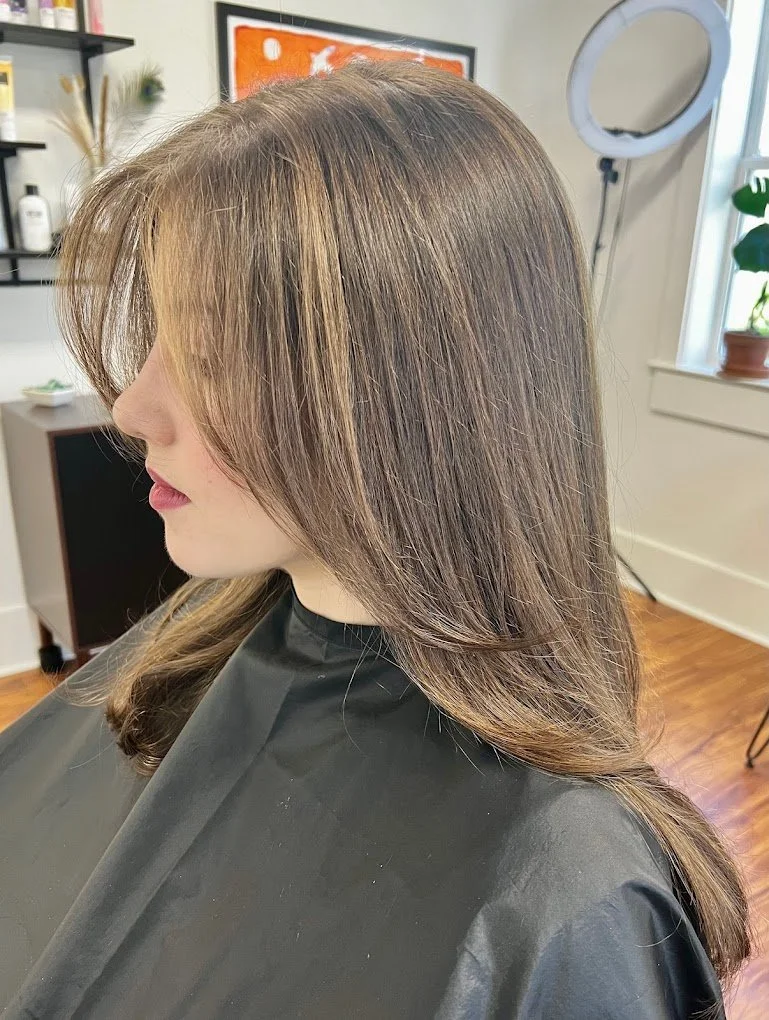 Side view of a woman with medium-length, layered brown hair with subtle highlights, sitting in a salon chair wearing a black cape. The salon has a ring light, a potted plant on a windowsill, and framed artwork on the wall.