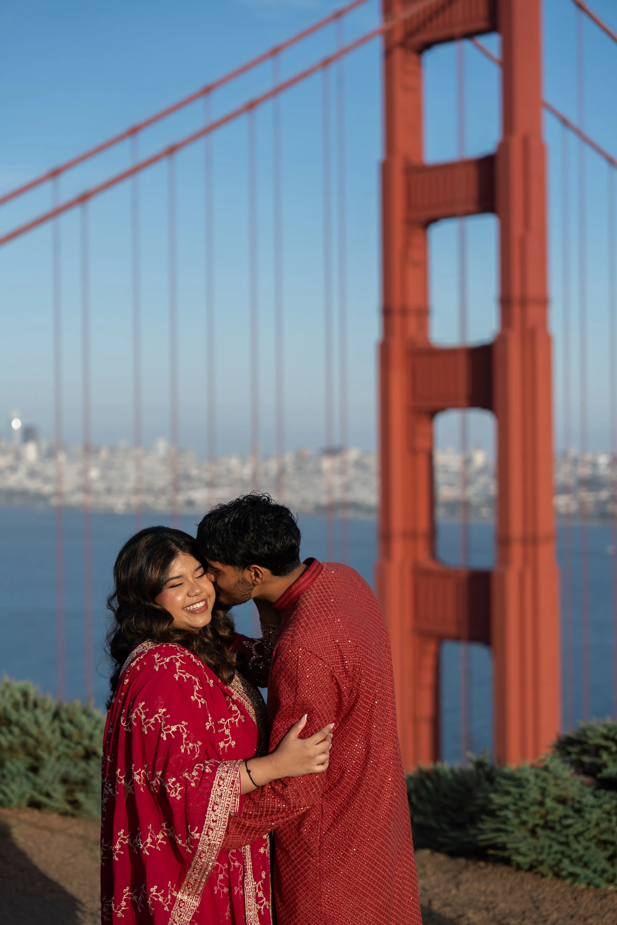 Fiancés kissing while taking engagement pre-wedding photos in San Francisco near Golden Gate Bridge wearing traditional Indian outfits