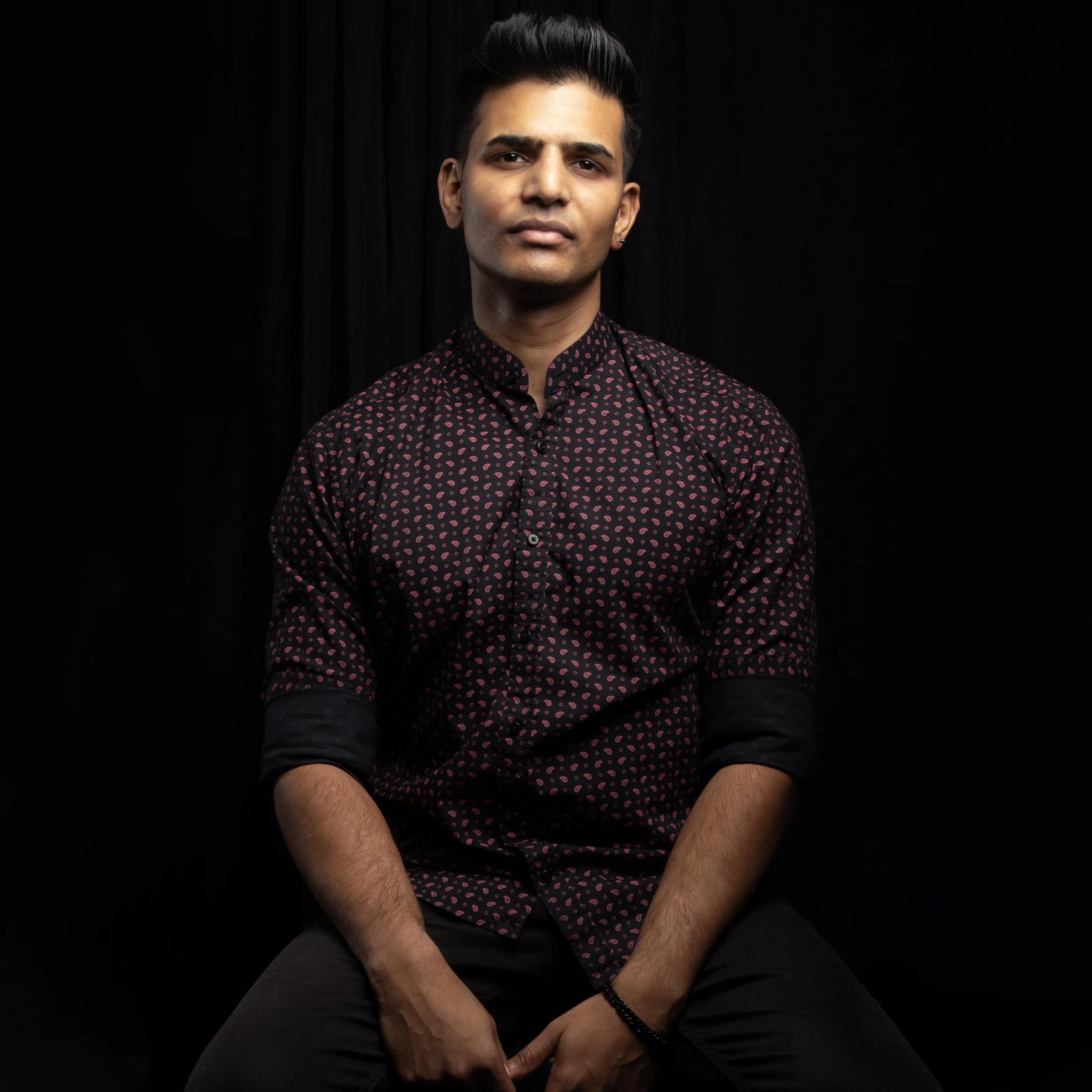 Portrait of a young man with dark hair styled upward, wearing a dark shirt with pink heart patterns, sitting against a black background.